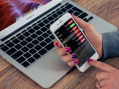 Image of a woman's hand using a phone in front of a computer with financial rankings.