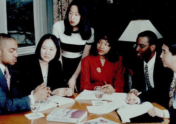 Six people in business attire sit around a table with books and paper in front of them.