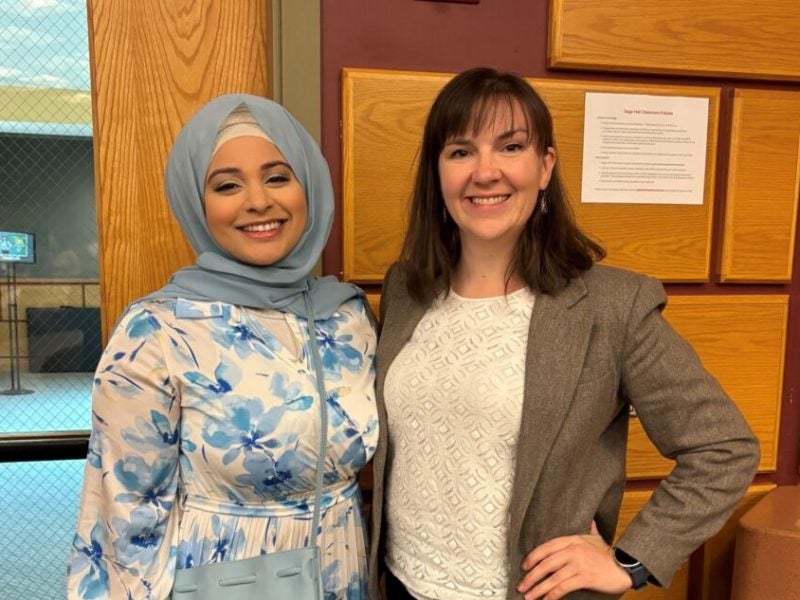 A young woman student posing in business casual attire alongside her professor outside a lecture hall