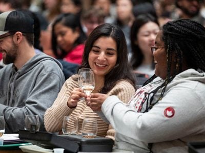 Students sitting next to each other in a crowded lecture hall smile and clink wine glasses before tasting the white wine in them.