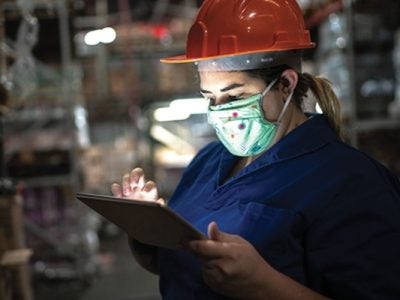 Person wearing a face mask and hardhat works on a touch screen in a warehouse.