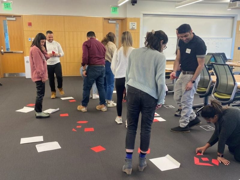Several Cornell MBA students stand over papers and look downward at small notes on the floor during a leadership workshop