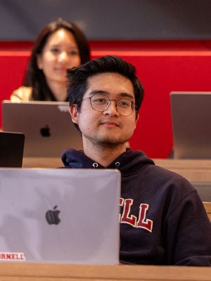 Student in a Cornell hoodie looking up from a laptop in a lecture hall.