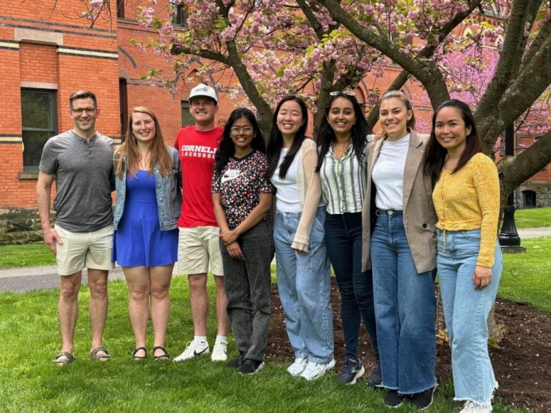 Young men and women in casual attire pose next to Sage Hall and a flowering tree as part of their roles as Grand Challenges coaches