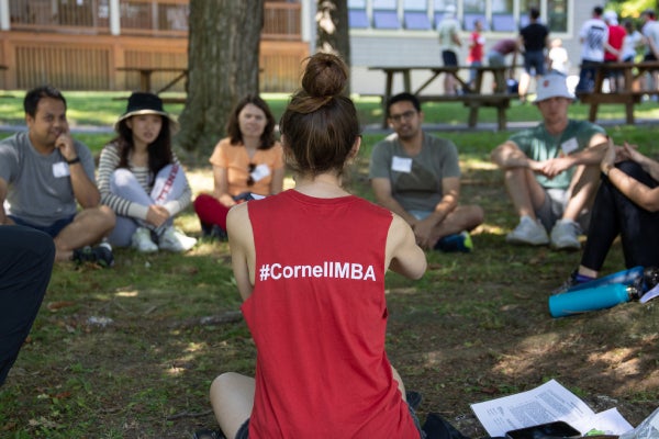 The back of a young woman sitting on the grass wearing a red tank top shirt with the hashtag #CornellMBA speaks to fellow Cornell MBA students as part of Johnson Outdoor Experience (J.O.E.)