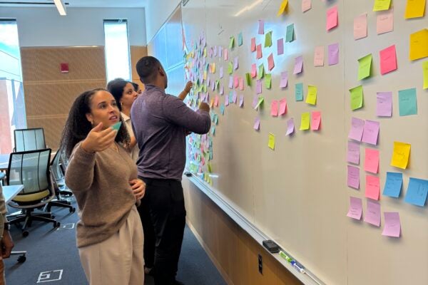 A young woman looks intently at a dry erase board full of brightly colored square sticky notes in front of her, looking for the spot to place her sticky note among other Cornell MBA students beside her