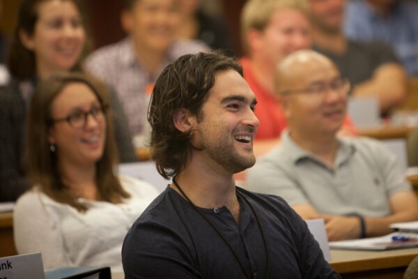 A young man in casual attire smiles during a lecture among other Cornell MBA students in a lecture hall