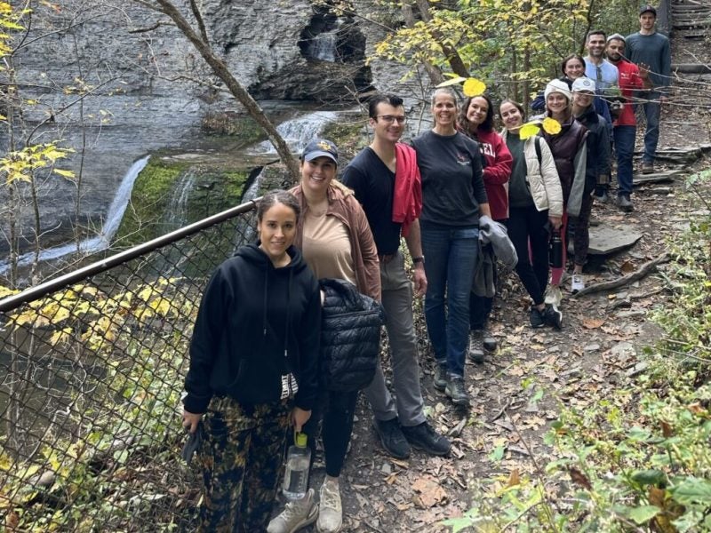 Young women and men pose outdoors in recreational attire against a black chain link fence and a waterfall as part of their Roy H. Park Leadership Fellowship program during their time as Cornell MBA students