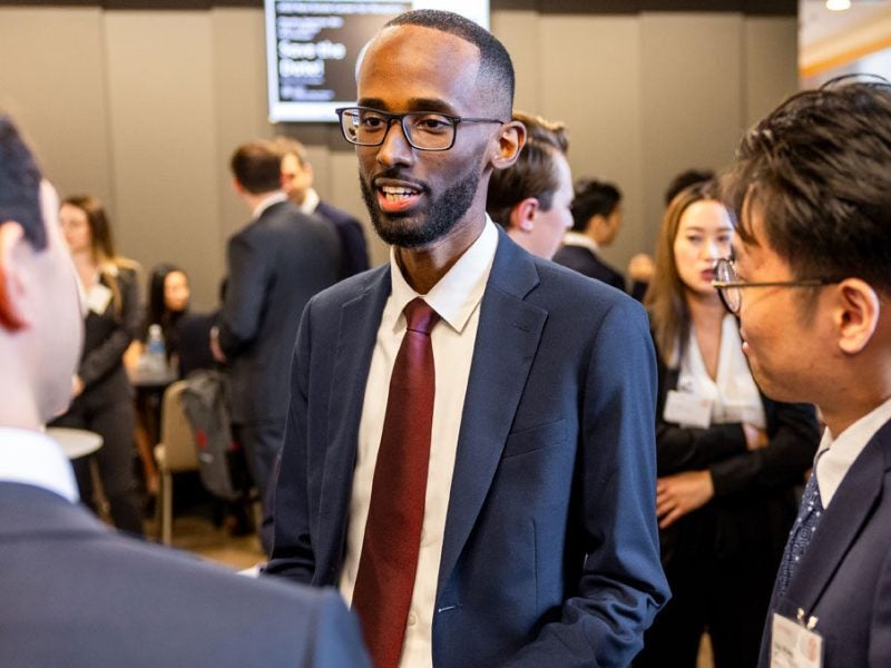 A young man in glasses and business attire speaks with fellow Cornell MBA students at a large social function
