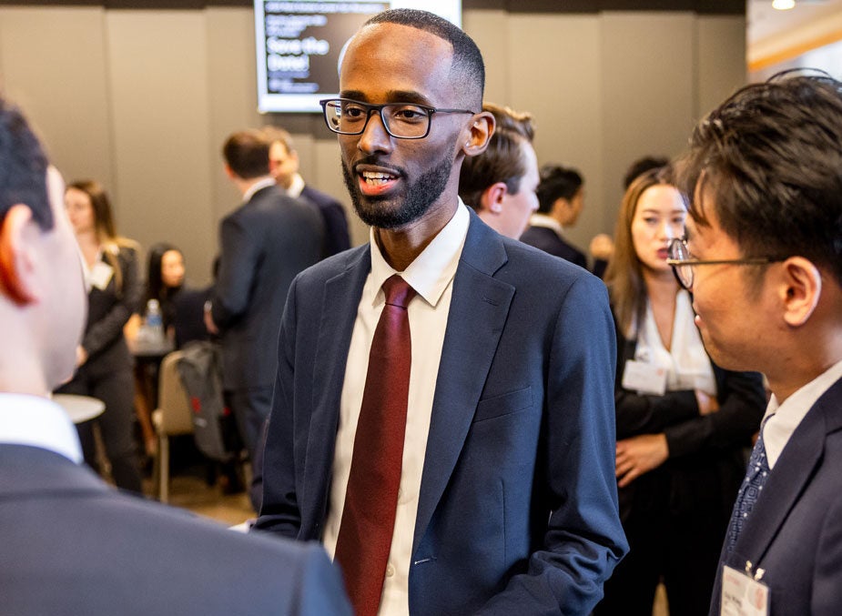 A young man in glasses and business attire speaks with fellow Cornell MBA students at a large social function