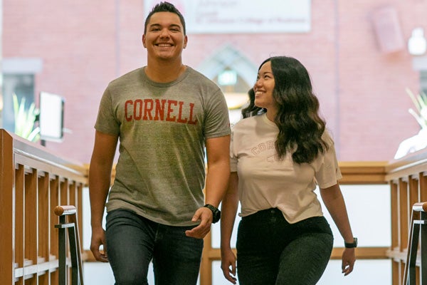 A young man on the left and a young woman on the right, both wearing Cornell t-shirts with jeans, walk up a staircase smiling in between classes as part of the Cornell MBA program
