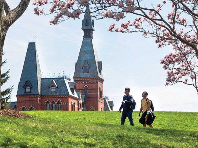 Two young men walk on a large grassy knoll with the upper portion of the Sage Hall gothic-inspired architecture building behind them on Cornell University's central campus