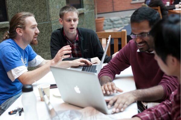 A team of young men workshop ideas at a table with a laptop as part of a project in the Cornell MBA leadership-focused curriculum