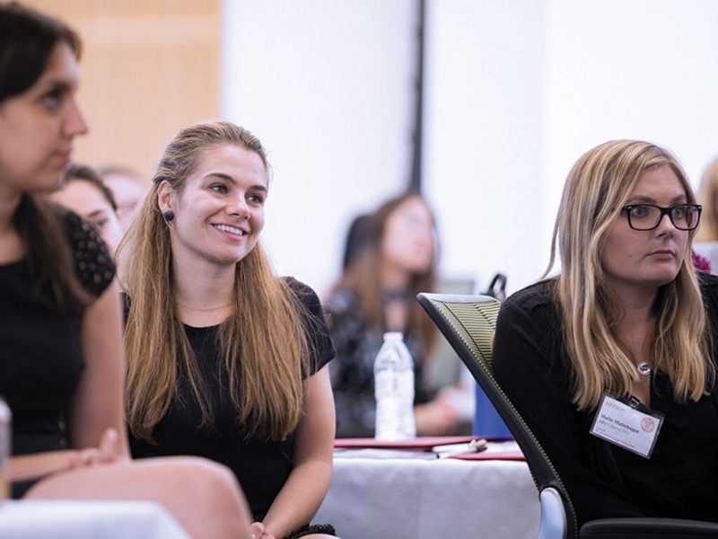Several young women in business attire sit at round tables in a well lit room, learning more about the Cornell MBA program