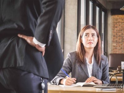 A woman wearing a suit sitting at a table looks up uncertainly at a person standing with hand on hip.