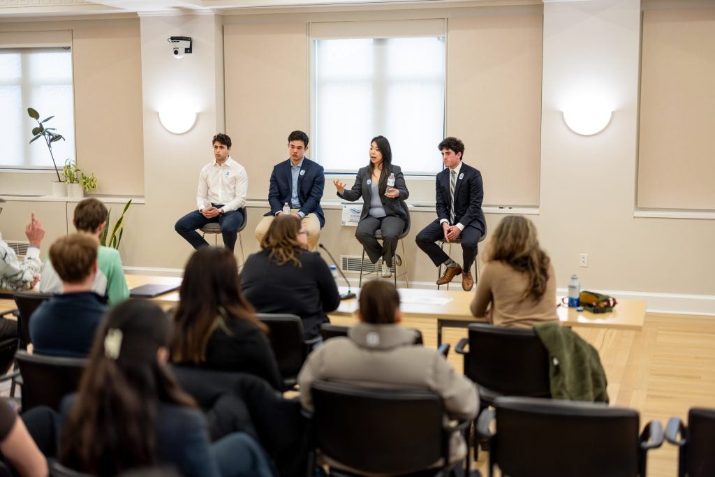 Four students in formal business attire sit on stools at the front of the room speaking to an audience.