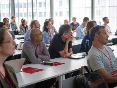 BDRM conference attendees sit and listen to a presentation with sunlight streaming in from a wall of windows.