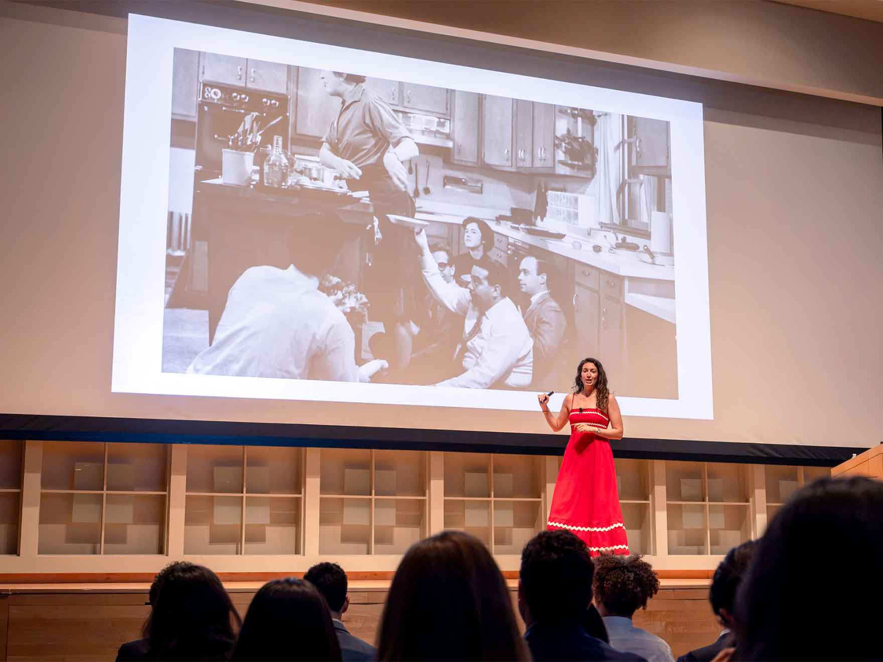 A woman in a red dress speaks on stage, a black-and-white image of a celebrity chef projected behind her.