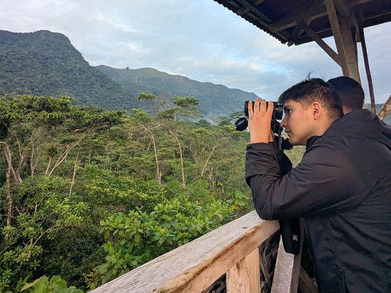 Student using binoculars to look out at the forest and mountains of Peru while on a trip for the Grand Challenges program.