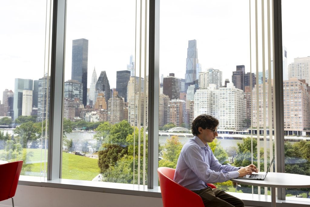 A Baker Program student sits working on his laptop with the Manhattan skyline visible through the wall of windows behind him.