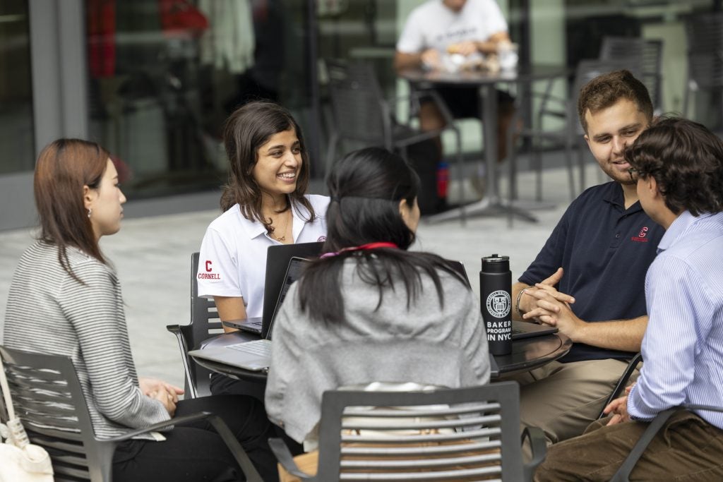 Five Baker Program students sit outside at a table with laptops open in front of them.
