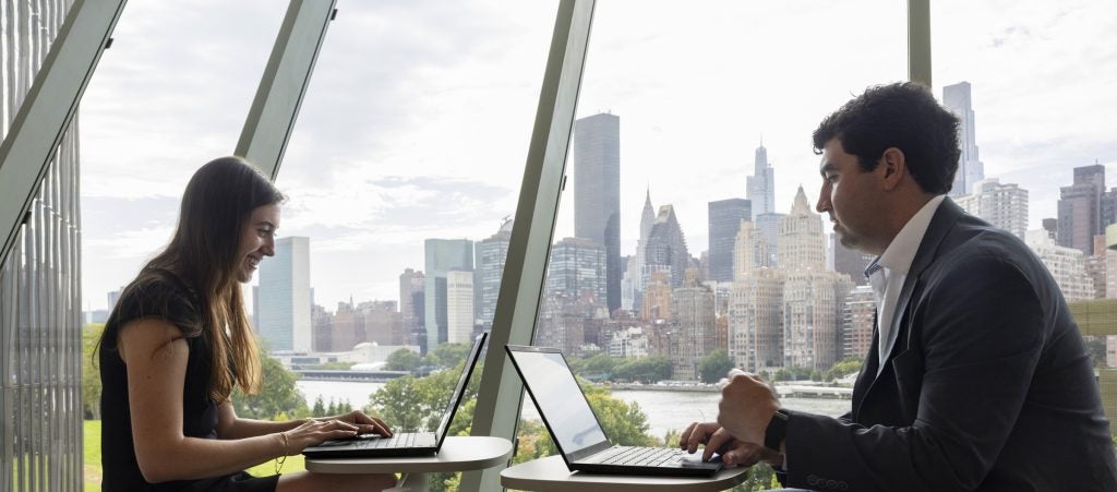 Two Baker Program students sit working on laptops in the Tata Innovation Center with the Manhattan skyline visible behind them.