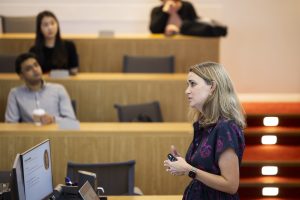 A professor stands in front of a tiered classroom speaking with a clicker in her hand.