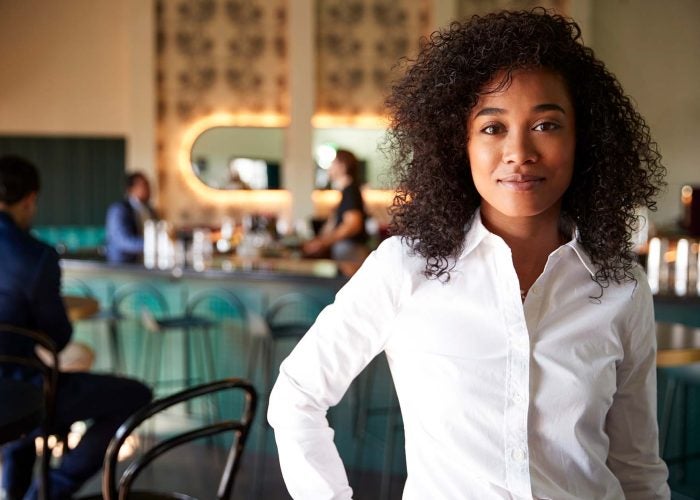 Black woman in white shirt stands in cocktail bar.