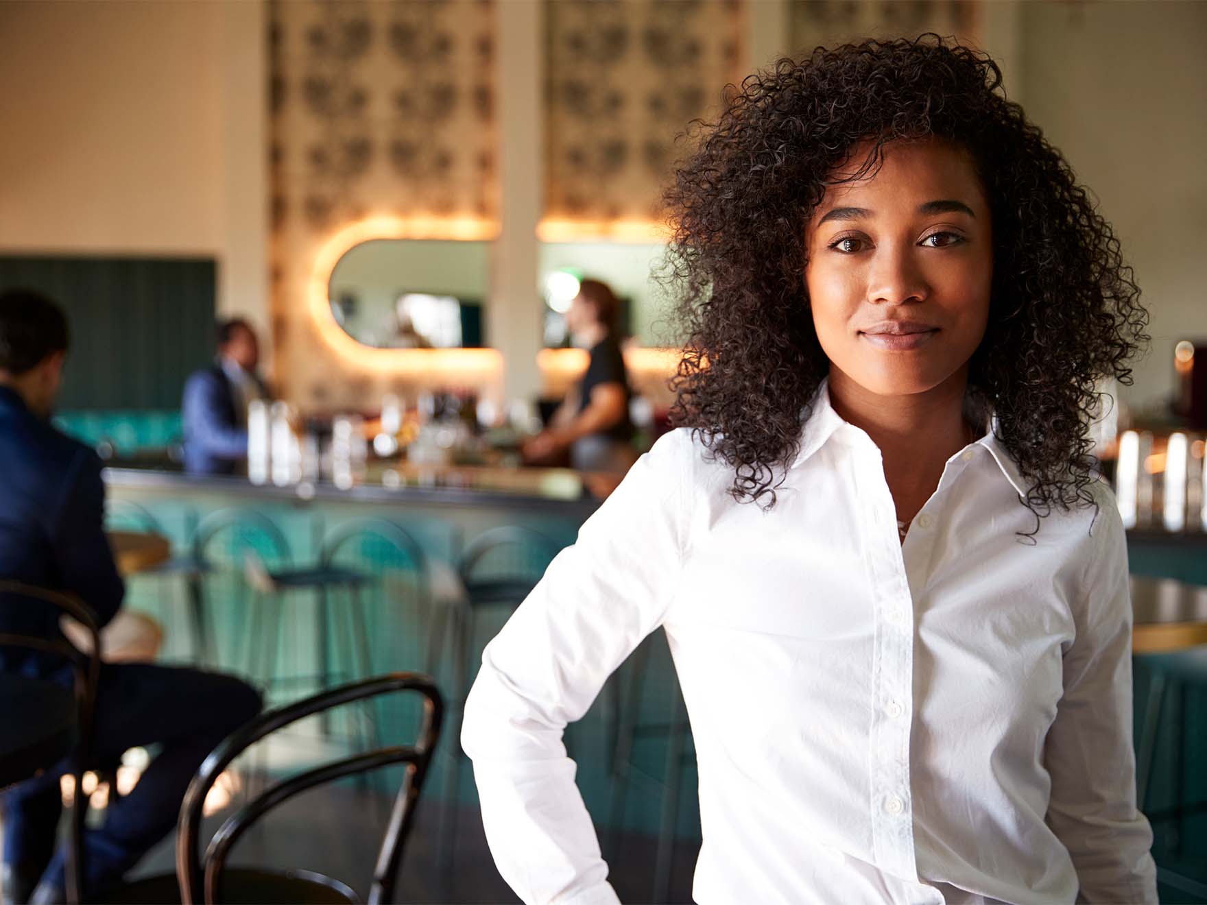 Black woman in white shirt stands in cocktail bar.