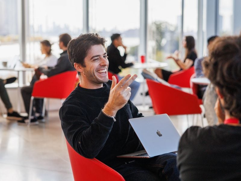 A young man enrolled in the Johnson Cornell Tech (JCT) MBA program smiles while working on a group project in cozy red chairs in a sunlight atrium collaboration space in Tata Innovation Center at Cornell Tech's campus on Roosevelt Island in New York City (NYC)
