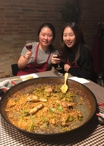 Photo of Nicole and a classmate holding wine behind a large bowl of paella