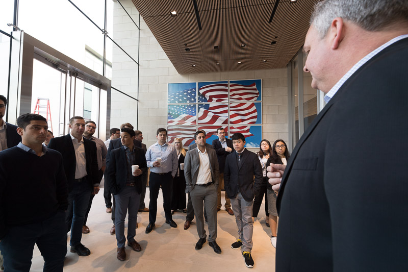 Photo of a group standing and listening to a speaker with an American flag in the background