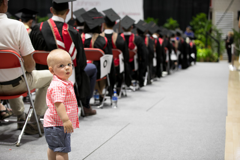 Photo of a baby standing in the aisle amongst graduates