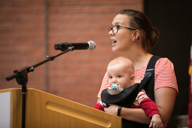 Photo of a women speaking at a podium holding a baby