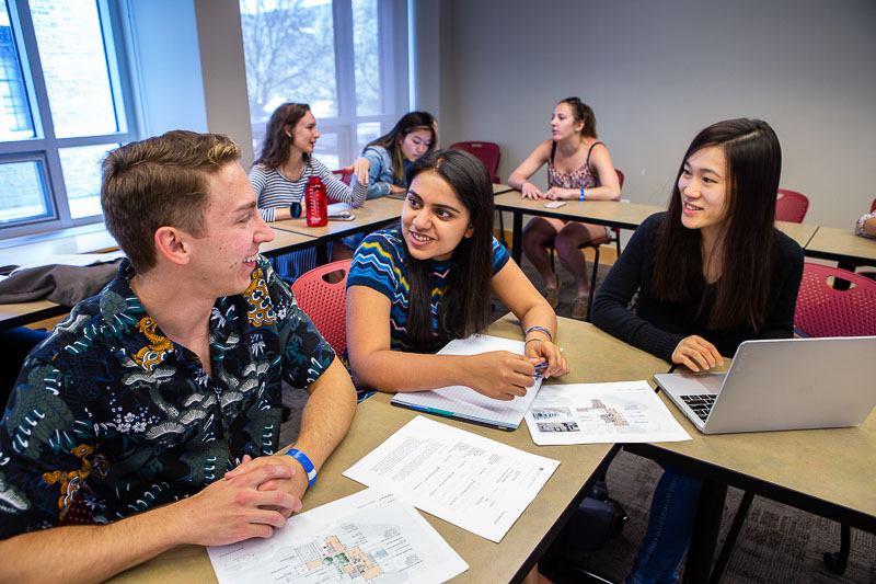 Photo of students sitting in a classroom; three are talking around papers