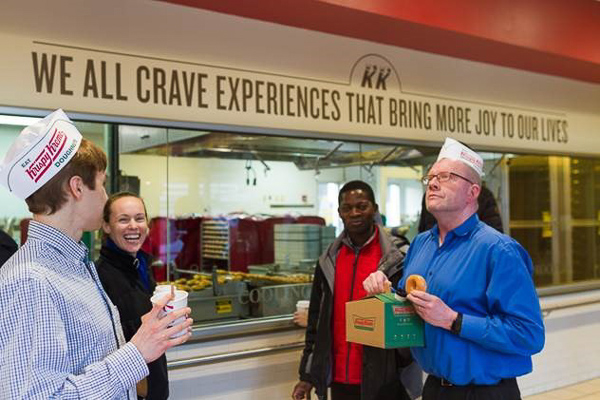 Photo of students and workers wearing Krispy Kreme hats in front of a sign that says "we all crave experiences that bring more joy to our lives"