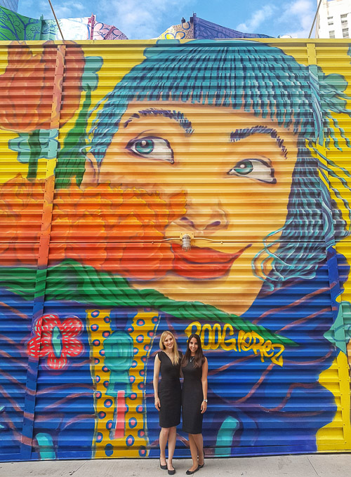 Two women standing in front of a colorful mural on a building