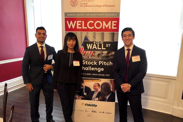 Three students stand in front of the MBA Stock Pitch Challenge welcome sign