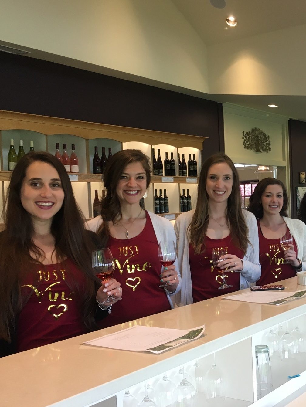 Four women wearing matching shirts and holding wine glasses