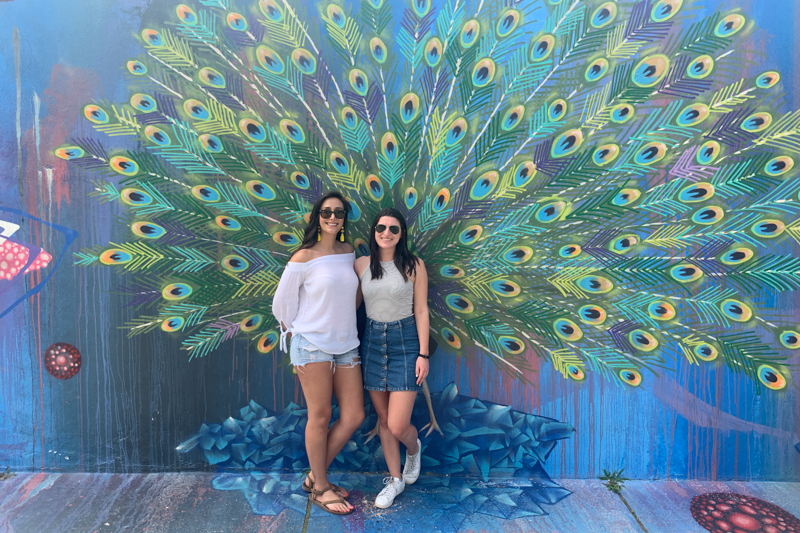 Two women standing in front of a peacock mural