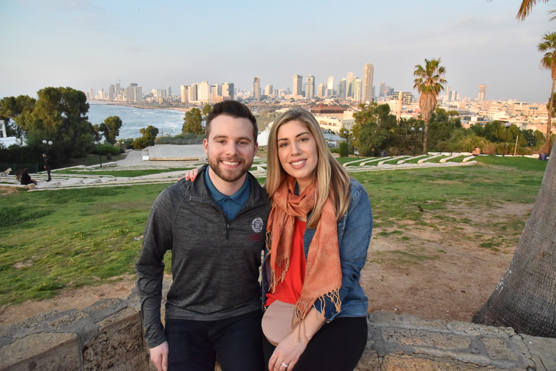 Nick and Katie posing for a photo in front of the Tel Aviv skyline and water