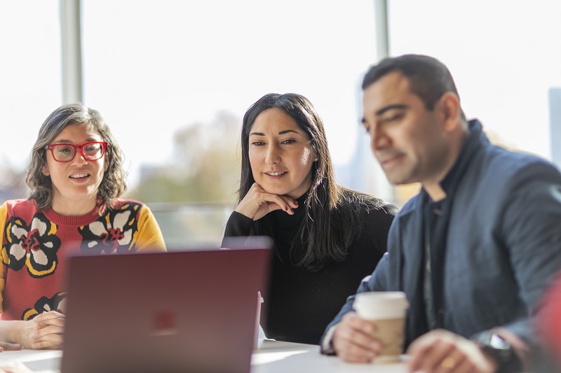 Three students sit together, viewing a laptop screen.