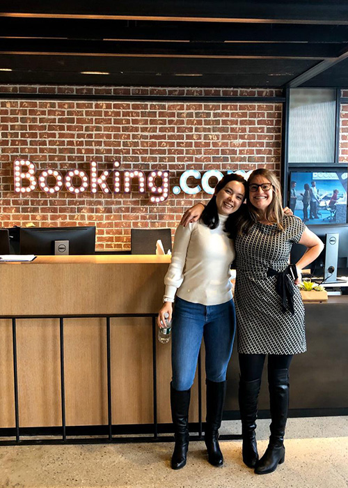 Emma Brett and Cassandra Martinez stand in front of the Booking.com company sign.