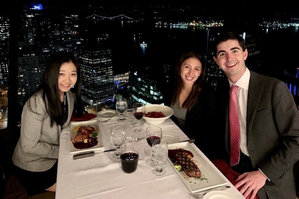 : Carol Wang, Tori Weissman, and Tim Bergin sit at restaurant table with Vancouver skyline at night behind them.