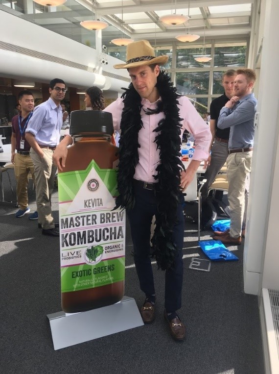 Taylor stands next to a cardboard Kombucha wearing a boa and cowboy hat