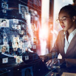 Business woman of color typing on a laptop with a hologram of a globe and connections to many headshots in the foreground