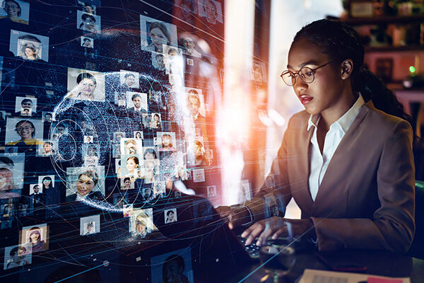 Business woman of color typing on a laptop with a hologram of a globe and connections to many headshots in the foreground
