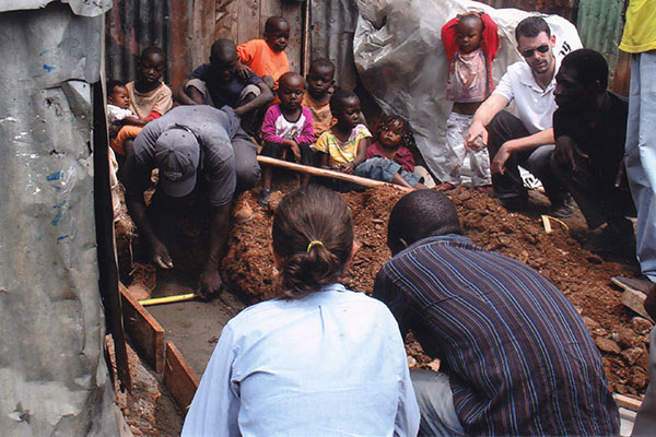 many children and several adults look on as men lay the foundation to install a bench