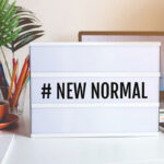 stock photo: “new normal” sign in the middle of a work desk with a cup of colored pencils, a plant, a laptop, and a cell phone