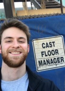 The author smiles for a selfie next to a sign that reads “Cast Floor Manager.”